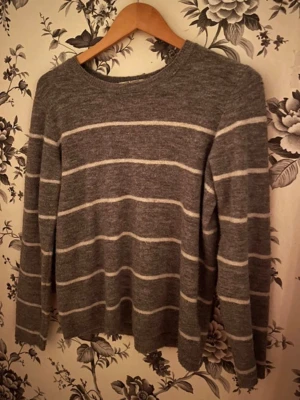 Grå randig tröja med rund hals - Mysig grå tröja med vita horisontella ränder och rund halsringning. Tröjan har långa ärmar och en avslappnad passform, perfekt för en chill vardagslook. Materialet ser ut att vara mjukt och stickat, vilket gör den skön att bära.
