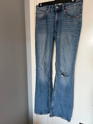 Bootcut jeans i ljusblå denim - Säljer ett par ljusblå bootcut jeans  med klassisk femficksmodell och slitning på ena benet. Jeansen har låg midja, raka ben som går ut något nedtill och är tillverkade i mjuk denim. Perfekta för en avslappnad och trendig look.