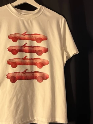 Vit t-shirt med röda bilar - Vit t-shirt i bomull med tryck av fyra röda cabrioletbilar på framsidan. Klassisk passform och rund halsringning. Perfekt för dig som gillar retrostil och coola motiv. Storlek L men passar för M & S. 