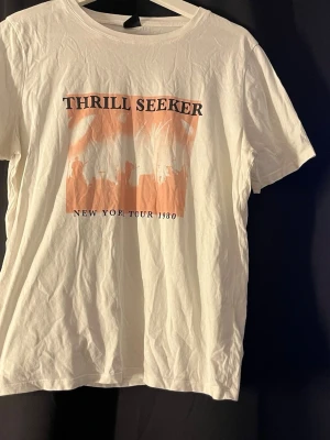 Vit THRILL SEEKER t-shirt - Vit t-shirt med tryck i orange och svart text 'THRILL SEEKER' och 'NEW YORK TOUR 1980' framtill. Klassisk rund hals och korta ärmar. Perfekt för dig som gillar retrovibbar och coola prints. Storlek L men är som en S