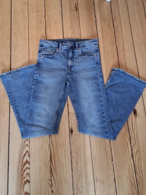 Blå utsvängda jeans med fem fickor - Säljer ett par klassiska blå jeans med utsvängda ben och fem fickor. Jeansen har bälteshällor, knappgylf och subtila slitningar för en avslappnad look. Perfekta till vardags och passar till det mesta. Materialet är denim i bomull.