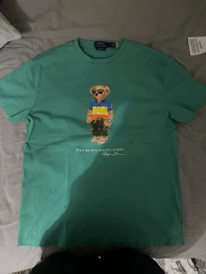 Grön Polo Bear t-shirt från Ralph Lauren - Snygg grön t-shirt från Polo Ralph Lauren med klassisk fit. På bröstet finns ett tryck av den ikoniska Polo Bear i färgglada kläder och solglasögon. T-shirten har rund hals och är tillverkad i mjuk bomull för skön känsla.