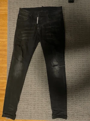 Svarta jeans från Dsquared. Äkta! - Säljer ett par svarta skinny jeans från D2 med slitningar på knäna och lätt tvättade detaljer. Jeansen har fem fickor, dragkedja och knapp framtill samt en patch med logga bak i midjan. Perfekta för dig som gillar en slim fit och streetstyle. Pris kan diskuteras