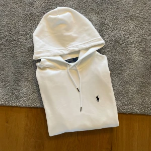 Vit hoodie från Polo Ralph Lauren - Säljer en stilren vit hoodie från Polo Ralph Lauren med klassisk huva och dragsnören. Tröjan har den ikoniska lilla svarta loggan broderad på bröstet och en känguruficka framtill. Perfekt för en clean och avslappnad look.