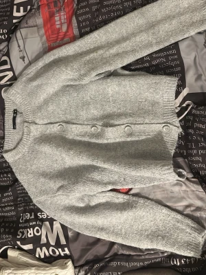 Ljusgrå stickad kofta från lager 157 - Mysig ljusgrå stickad kofta från lager 157 med rund halsringning och stora vita knappar framtill. Koftan har långa ärmar och ribbade muddar vid ärmslut och nederkant. Perfekt för lager på lager och enkel att matcha.