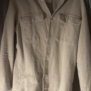 Beige overshirt - Säljer en stilren beige overshirt  med två bröstfickor och knappar framtill. Skjortan har klassisk krage och är tillverkad i ett mjukt material som passar perfekt till lager-på-lager-stil.