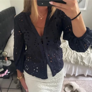 Svart blus med hålmönster och knappar - Säljer en svart blus med hålmönster och knappar framtill. Blusen har v-ringning och långa ärmar med lätt puff. Perfekt att styla med kjol eller jeans för en trendig look.