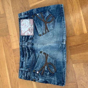 Blå jeanskjol från Primigius Brand - Säljer en cool blå jeanskjol med slitningar och dekorativa bruna sömmar på fickorna. Kjolen har två framfickor och två bakfickor med snygga detaljer. Perfekt för dig som gillar en avslappnad och trendig stil.