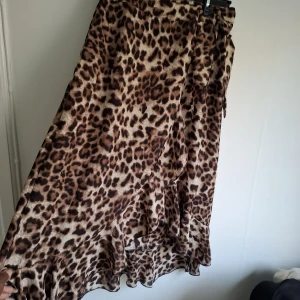 Leopardmönstrad omlottkjol med volang - Säljer en snygg leopardmönstrad omlottkjol med volangkant. Kjolen har en längre modell och knyts i midjan för en justerbar passform. Perfekt för dig som vill sticka ut med ett djurmönster.