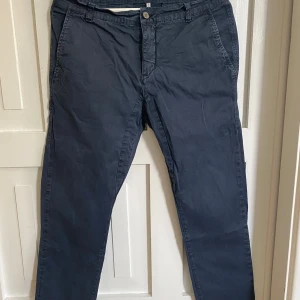 Mörkblå chinos från Scout - Snygga mörkblå chinos från Scout med klassisk passform. Byxorna har snedställda fickor fram, två bakfickor med knapp och stängs med dragkedja och knapp. Perfekta för en stilren look.