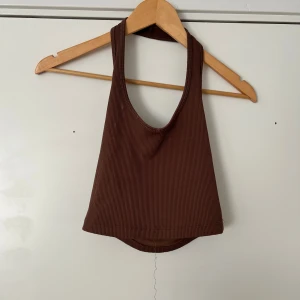 Brun ribbad croppad linnetopp - Säljer en brun ribbad croppad linnetopp med djup urringning och breda axelband. Perfekt för varma dagar eller som ett lager under skjortor och jackor. Enkel och stilren design.