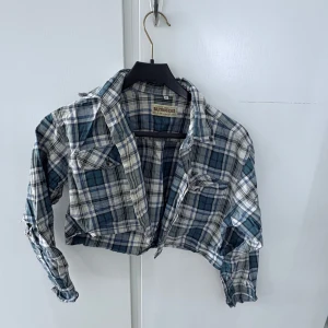 Blå och vit rutig croppad skjorta från Eastbourne - Säljer en croppad rutig skjorta från Eastbourne i blått, vitt och svart. Skjortan har långa ärmar, klassisk krage och två bröstfickor med knappar. Perfekt för en avslappnad och cool stil.