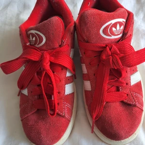 Röda Adidas Campus sneakers - Säljer ett par röda Adidas Campus sneakers med vita ränder och logga på både plös och häl. Skorna har klassisk låg modell, snörning och ovandel i mocka. Yttersulan är i gummi och har ett mönster för bra grepp. Perfekta för en sportig och stilren look.