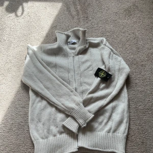 Vit stickad ziptröja från Stone Island - Säljer en vit stickad tröja från Stone Island med dragkedja och hög krage. Tröjan har ribbade muddar och nederkant samt den klassiska Stone Island-loggan på ärmen. Perfekt för kyligare dagar. Pris går att förhandla vid snabb affär!!⭐️⭐️