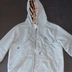 Grå hoodie med dragkedja från Burberry - Säljer en grå hoodie från Burberry med klassiskt rutigt foder i huvan. Tröjan har dragkedja framtill, två fickor och justerbar huva med snören. Perfekt för en stilren och avslappnad look.