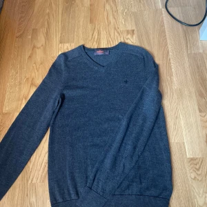 Morris tröja merinoull - Säljer en mörkgrå stickad tröja från Morris med v-ringad hals och diskret broderad logga på bröstet. Tröjan har långa ärmar och ribbade muddar vid ärmslut och nederkant. Perfekt för lager-på-lager och stilren look. Den har ett lite märke efter lagning som man ser på en av bilderna men den syns nästan inte i verkligheten. Är i storlek S men passar även XS