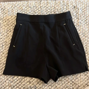 Svarta shorts med gulddetaljer - Stilrena svarta shorts med hög midja och snygga gulddetaljer vid fickor och knäppning. Klassisk passform med raka ben och diskreta fickor både fram och bak. Perfekta för en dressad look.