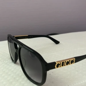 Svarta solglasögon från Gucci - Snygga svarta solglasögon från Gucci med klassisk pilotform och mörka glas. Bågarna är i svart plast med guldiga Gucci-detaljer på sidorna. Perfekt accessoar för dig som vill sticka ut med en lyxig touch.