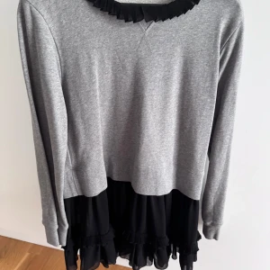 Grå och svart klänning från MM6 Maison Margiela - Supersnygg klänning från MM6 Maison Margiela med grå sweatshirt-överdel och svart volangkjol. Långärmad modell med rund hals och volangdetaljer nertill. Perfekt för dig som gillar en mix av sportigt och feminint.