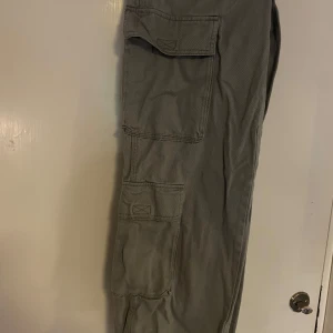 Grå cargobyxor med stora fickor - Säljer ett par grå cargobyxor med flera stora fickor på benen och klassisk cargostil. Byxorna har normal passform och är perfekta för dig som gillar praktiska detaljer och en avslappnad look.