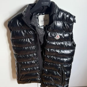 Svart dunväst från Moncler - Säljer en svart, glansig dunväst från Moncler med dragkedja och klassisk Moncler-logga på bröstet. Västen har två fickor med dragkedja och quiltad design. Perfekt för lager-på-lager-stil under kyliga dagar.