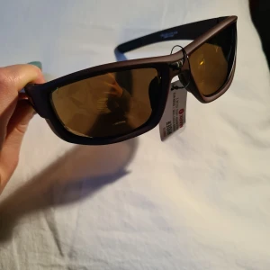 Bruna sportiga solglasögon från Bliz - Coola solglasögon från Bliz med bruna bågar och gulaktiga spegelglas, med riktigt bra solskydd. Modellen heter Rider och har en sportig design med breda skalmar och Bliz-logga i guld på sidan. Perfekta för utomhusaktiviteter. Helt nya i originalförpackning!