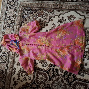 Rosa blommig klänning med knappar - Säljer en rosa klänning med blommigt mönster i orange, grönt och vitt. Klänningen har korta puffärmar och knappar framtill. Perfekt för sommarens tillfällen och har en feminin, lätt utställd passform.