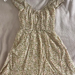 Blommig kortklänning från NA-KD - Söt blommig klänning från NA-KD i ljusa toner med grönt och rosa mönster. Klänningen har korta ärmar, smock i midjan och knytning vid halsen!! 