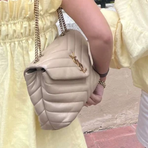 Beige Saint Laurent (intressekoll) - Säljer en beige quiltad axelväska från Saint Laurent med guldfärgad kedja och YSL-logga framtill. Väskan har en klassisk v-formad design och är tillverkad i skinn. Perfekt för dig som vill ha en stilren och lyxig accessoar.  (Ältihetsbevis kommer med köpet) 