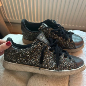 Glittriga sneakers från Victoria - Säljer ett par svarta sneakers från Victoria med glittrigt mönster över hela skon. Skorna har svarta snören, rund tå och silvriga detaljer på tån och hälen. Perfekta för dig som vill sticka ut lite extra!
