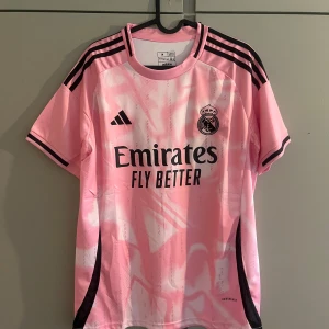 Real Madrid rosa fotbollströja från Adidas - Säljer en rosa Real Madrid fotbollströja från Adidas med svarta detaljer och klubbmärke på bröstet. Tröjan har korta ärmar, svart Adidas-logga och sponsortryck på framsidan. Tillverkad i lätt och ventilerande material.