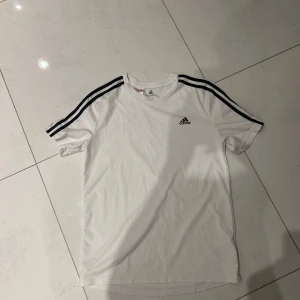 Vit t-shirt från Adidas med svarta ränder - Vit t-shirt från Adidas med klassiska svarta ränder längs axlarna och Adidas-logga på bröstet. T-shirten har rund hals och är kortärmad, perfekt för sport eller vardag.