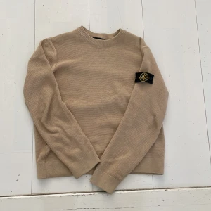 Beige stickad tröja från Stone Island - Säljer en beige stickad tröja från Stone Island med klassisk patch på ärmen. Tröjan har rund halsringning och långa ärmar. Perfekt för lager-på-lager och stilren vardagslook. Har ett par fläckar som knappt syns. Storlek 152-158