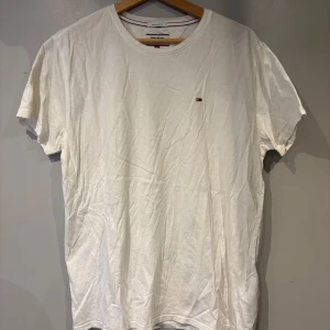 Vit t-shirt från Tommy Hilfiger - Vit t-shirt från Tommy Hilfiger med liten broderad logga på bröstet. Klassisk rund halsringning och normal passform. Perfekt basic-plagg med diskret branding och mjukt bomullsmaterial.