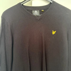 Svart v-ringad tröja från Lyle & Scott - Svart långärmad tröja från Lyle & Scott med v-ringning och den klassiska gula loggan broderad på bröstet. Enkel och stilren design som passar perfekt till jeans eller chinos.