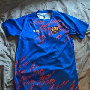 FC Barcelona blå fotbollströja - Säljer en blå FC Barcelona fotbollströja med röd grafiskt mönster och klubbmärke på bröstet. Tröjan har korta ärmar och rund hals. Perfekt för dig som vill visa ditt stöd för Barca på planen eller läktaren.