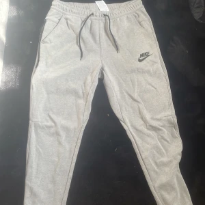 Grå Nike tech byxor  - Snygga grå mjukisbyxor från Nike med svart logga på vänster sida. Byxorna har dragsko i midjan och sidofickor, perfekta för chill eller träning. Tillverkade i mjukt material för extra komfort.