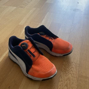 Orange och marinblå golfskor från Puma  - Säljer ett par sportiga sneakers från Puma i orange och marinblått med vit sula och svart yttersula. Skorna har rund tå, platt sula och en unik snörningslösning med snabbstängning. Perfekta för träning eller vardag.
