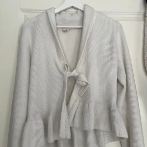 Vit ribbad kofta med knytband och volang - Mysig vit kofta i ribbat material med huva, knytband framtill och volangdetalj nedtill. Perfekt för lager-på-lager och en avslappnad stil. Säljes då den inte längre kommer till användning.