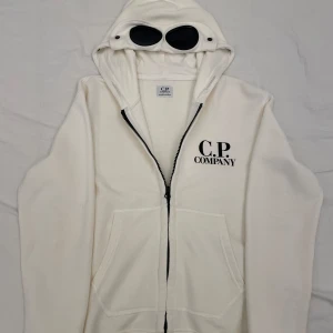 Vit zip hoodie med goggles från C.P. Company - Vit zip hoodie från C.P. Company med svarta goggles i huvan och svart dragkedja. Klassisk tryckt på bröstet och två stora fickor framtill. Aldrig använd! Nypris 2295, mitt pris 600! Såklart 100% äkta!✅Skulle säga att den passar mellan 155-160! Ställ gärna frågor om ni har några funderingar!! Priset är inte hugget i sten 🔨🪨!
