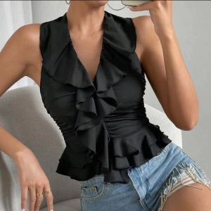 Svart volangblus med knyt i nacken - Snygg svart blus med djup v-ringning och volangdetaljer framtill. Toppen har en omlottkänsla och knyts med ett brett band i nacken. 