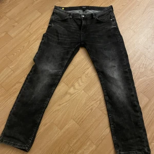 Svarta jeans från SMOG - Säljer ett par svarta jeans från SMOG med klassisk femficksdesign och knappgylf. Jeansen har en lätt tvättad look och raka ben. Perfekta för dig som gillar en enkel och stilren stil.