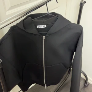 Svart hoodie med dragkedja från Weekday - Säljer en svart hoodie från Weekday med dragkedja framtill och huva. Tröjan har långa ärmar och två fickor framtill. Perfekt för en avslappnad stil.