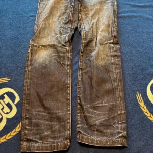 Grå slitna jeans med dragkedjedetalj - Säljer ett par grå jeans med snyggt sliten tvätt och dragkedja vid ena fickan fram. Byxorna har raka ben och klassiska bakfickor med dekorativa sömmar. Perfekta för en avslappnad och cool stil.