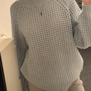 Ljusblå stickad tröja från 157 - Mysig ljusblå stickad tröja från 157 med grovstickat mönster och hög krage. Tröjan har långa ärmar och en avslappnad passform, perfekt för kyliga dagar.