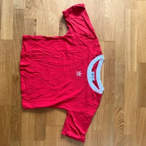 Röd t-shirt från Adidas med vit krage - Säljer en röd t-shirt från Adidas med vit krage och Adidas-logga broderad på bröstet. Klassisk och sportig stil, perfekt för dig som gillar streetwear. T-shirten har korta ärmar och är gjord i mjuk bomull.