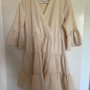Beige omlottklänning med volang - Säljer en beige omlottklänning med v-ringning och volangdetaljer på ärmar och kjol. Klänningen har trekvartsärm och en luftig passform, pris kan diskuteras.