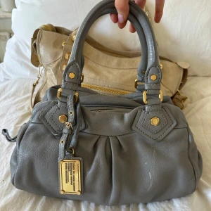 Ljusgrå Q Classic Baby Groove väska! - Snygg ljusgrå handväska i vintage skick från Marc by Marc Jacobs i äkta läder med gulddetaljer och dubbla handtag. Finns en repa på framsidan som syns på bilderna. Även lite färg som lossnat på underdelen av ena fästet för handtaget (syns också på bilden). I övrigt en välbevarad vintage modell! Långa bandet tillkommer även! Perfekt för att ge en stilren touch till din outfit. Skriv privat för fler bilder. Mått 34x18x12 (undersida). Kan hämtas hemma hos mig i Nacka eller skickas🤍