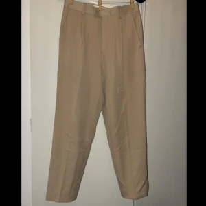 Beiga kostymbyxor  - Beiga kostymbyxor med pressveck. Byxorna har normal passform, hällor för bälte och diskreta fickor bak. Perfekta för en stilren look och passar till skjorta eller t-shirt.