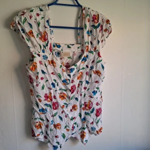 Vit blommig blus med knappar - Söt vit blus med färgglatt blommönster i rött, orange, blått och grönt. Blusen har korta ärmar, v-ringning och knappar framtill. Perfekt för en somrig look och lätt att matcha med jeans eller kjol.